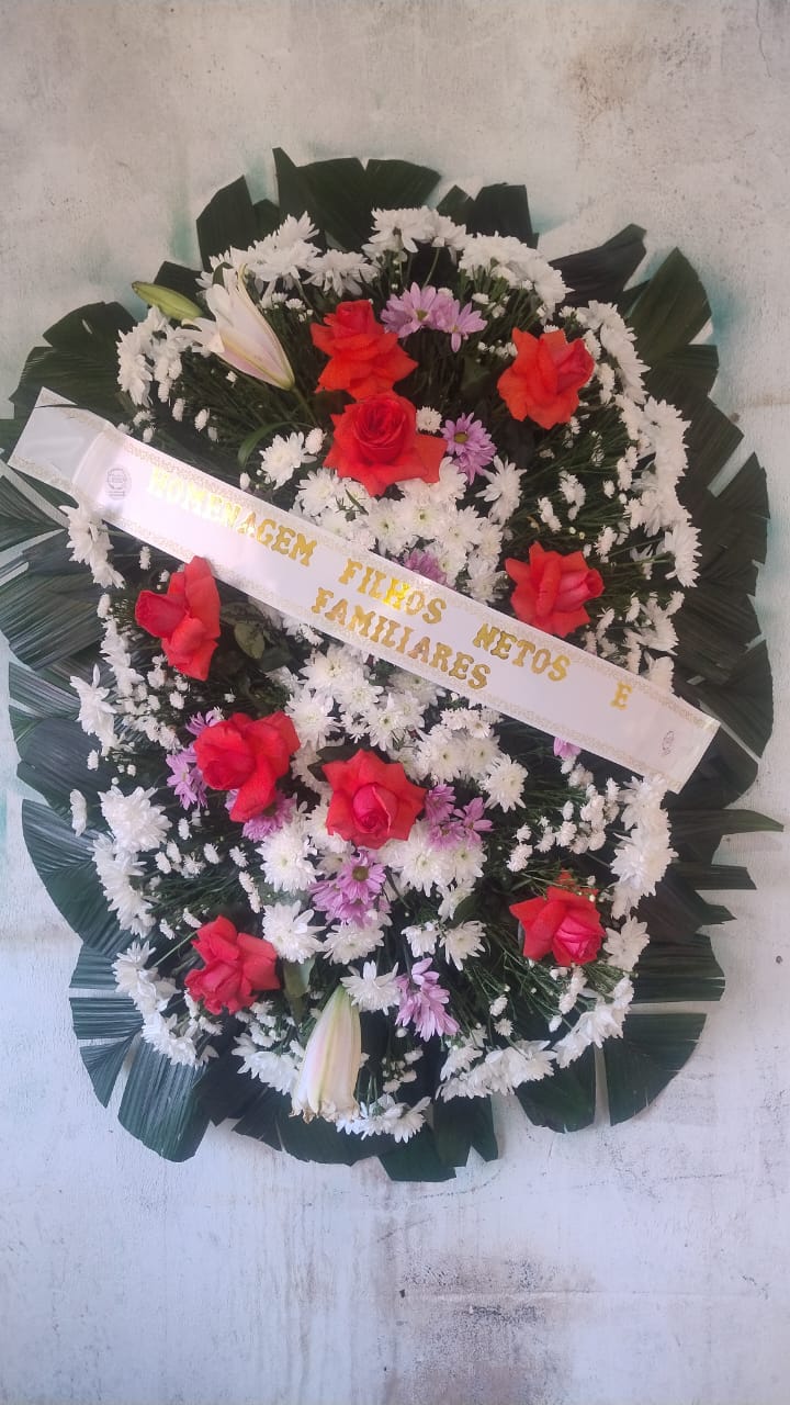arranjo fúnebre circular com rosas vermelhas, margaridas brancas e roxas sobre folhas de palma, com faixa bran