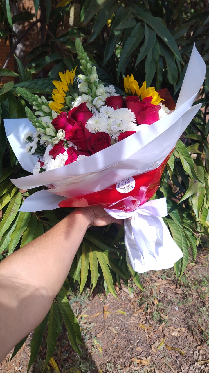 mão segurando um buquê de rosas vermelhas, margaridas brancas e girassóis com embalagem branca e vermelha e la