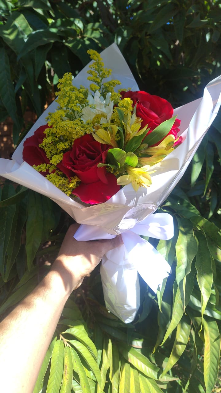 mão segurando um buquê de rosas vermelhas com flores amarelas e brancas embrulhado em papel branco com laço, d