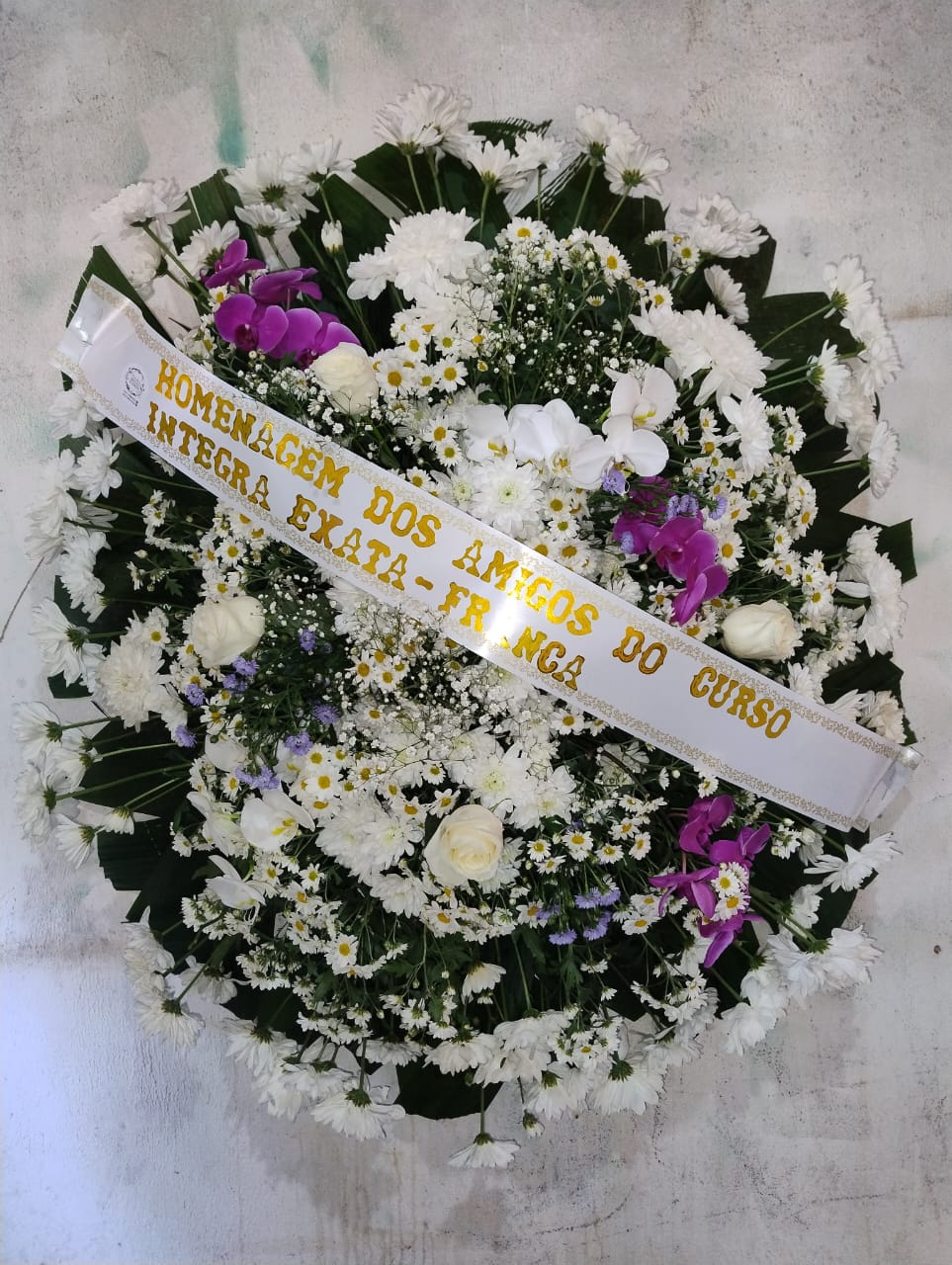 coroa de flores branca com margaridas, orquídeas roxas e rosas brancas com faixa dourada de homenagem