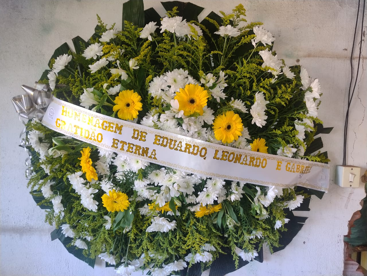 coroa de flores funerária com margaridas brancas e gerberas amarelas e faixa com dedicatória pendurada em parede clara